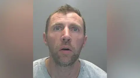 Cambridgeshire Police Samuel Jackson is in the middle of the image and has short brown hair and a beard. He is wearing a grey T-shirt and is sitting in front of a grey background.