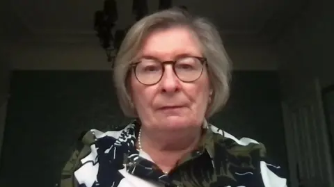 A representation   of Andrea Nelson, who has abbreviated  grey hair. She is wearing heavy   acheronian  rimmed glasses, a pearl necklace and a apical  with olive green, achromatic  and achromatic  floral patterns connected  it. 