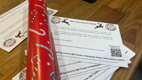 The image shows a handful of gift request forms on a wooden table with red Christmas wrapping paper.