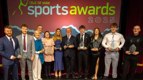 VANNIN PHOTOS The winners of the sports awards, who are all dressed smartly, lined up in a row holding their trophies in front of red screen that says Isle of Man Sports Awards 2025 in white and yellow lettering.