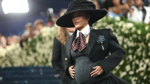 Reuters Rihanna appears at the 2025 Met Gala visibly pregnant. She is wearing a black and grey tailored outfit and large brim hat.