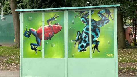 Nue/Katie Scott Two tree frogs painted on a bus shelter. The one on the left is red and blue and the one on the right is light blue with black spots and markings. 