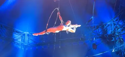 Laura is performing the splits through the hoop which is high up in the big to tent. She is smiling.