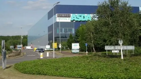 Robin Stott/Geograph A building, with an Asda sign on it, and a George sign. There is a roundabout outside the building, with greenery on it with signs. There are trees to the right and a road into the building, with cars on it and more signs. 