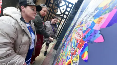 Middlesbrough Council Three woman take a closer look at the artwork. The feathers are brightly coloured and some text can be made out including one that reads: "Black lives matter". The wings are placed on a purple board.