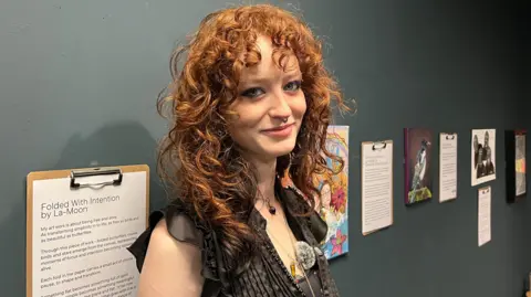 Sinead Diamond standing next to her artwork at Tullie. She has red curly hair down to her shoulders and a fringe. She has blue eyes and is wearing a black blouse. She has her back to a grey wall with artwork displayed and she is looking at the camera and smiling