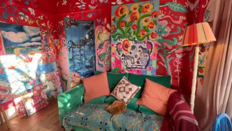 Alun Callender The picture shows a dark emerald green sofa with a ginger cat on it. The walls of the room are red with flowers painted on them. A large painting of flowers also leans against the wall.