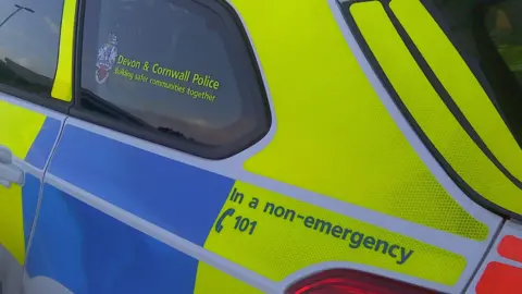 The side of a police car, which is flourescent yello and blue. The back window says 'Devon and Cornwall Police, building safer communities together'. Writing above the brake light says 'In a non-emergency call 101'.