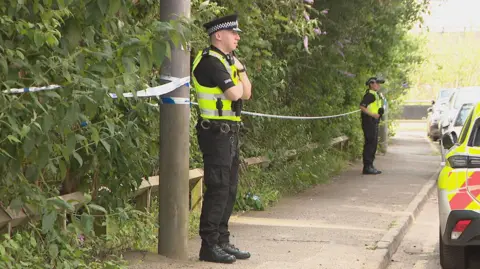 Two police officers standing on a pavement in front of a wooded area that has been cordoned off with blue and white police tape. There is a police car among vehicles parked on the road in front of them.