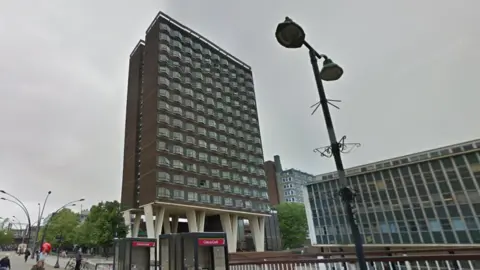 A 14-storey block of flats in Basildon, the building is made of house bricks with V-shaped white supporting piers at ground level. Each floor has 10 windows on the front face. There are several other modern buildings around it, and some old phone boxes in the foreground.