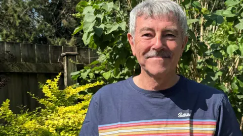 John Houghton John is outside standing on a garden. He is wearing a blue tshirt with coloured stripes