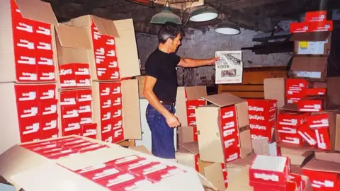 Jake Shoes An old photograph of a man in a storage area surrounded by piles of red Kickers shoe boxes.