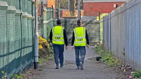 CST Two men wearing black caps, high-viz waistcoats and dark trousers walk away from the camera down a wide footpath, with high fences either side.