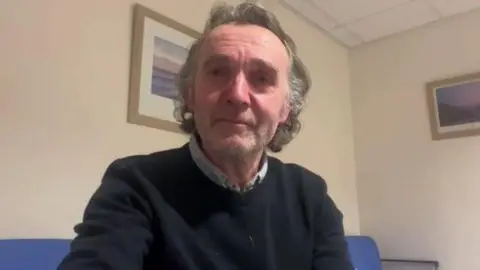 A man sitting down with dark grey hair and facial stubble. He is wearing a dark navy jumper with a shirt underneath.