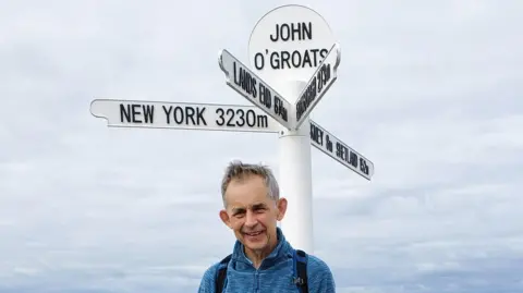 Mark Saunders Prof Saunders stands below a white and black John O'Groats sign which has arrows pointing to New York, Land's End and Orkney and Shetland. He has grey hair and is wearing a blue jumper. 