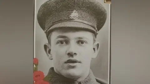 Supplied A black and white image of George Bell in a military uniform and hat. He is looking at the camera and his mouth is slightly open. There are red poppies over laid on the bottom left hand corner of the picture.