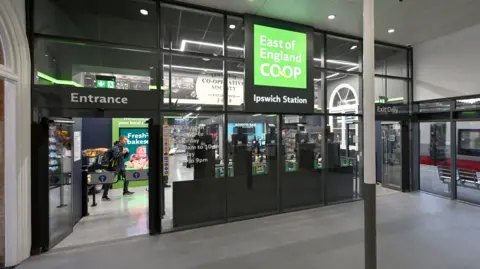 East of England Co-op The glass front of a shop. An illuminated green square sign says East of England Co-op in white, and Ipswich Station in white on a black background.