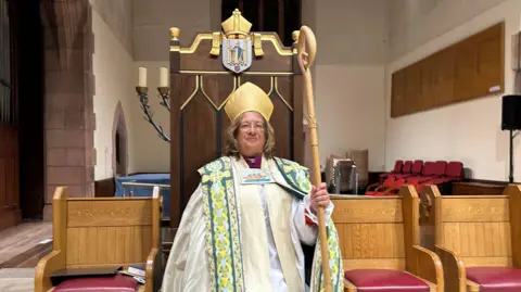 Tricia Hillas, she wears a decorated white robe and golf mitre and holds a gold staff while standing in front of a grand wooden chair.