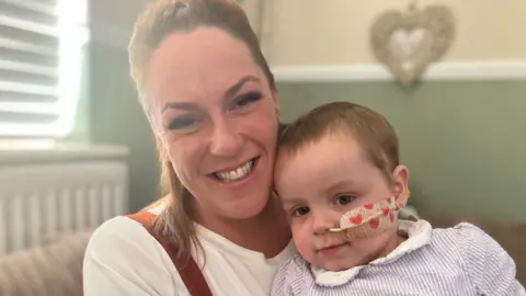 An image of Gemma holding her daughter Nancy on her lap while sitting on a sofa. Nancy has a thing tube attached to her nose with is stuck to her face with a heart-printed plaster. 
