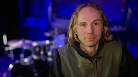 Martin Giles/BBC Ben Street looks directly at the camera. He has brown eyes and shoulder-length hair with a centre parting and a tattoo of a flower on his neck. He is wearing an olive green zip-up jacket. Behind him, blurred, is a drum kit bathed in blue light.