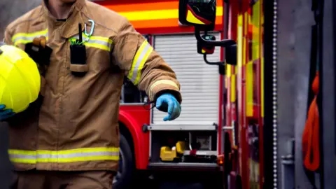 A stock image of a firefighter as they stand near two fire engines. The person is depicted from the shoulders down and is wearing a brown firefighter suit with reflective stripes. They are also carrying a neon yellow helmet.