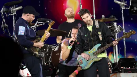 Coldplay is performing on stage at the Rose Bowl Stadium in Pasadena, California. Will Champion is towards the back playing the drums, to the left is guitarist Johnny Buckland, singer Chris Martin stands in the centre and bass player Guy Berryman is to the right. 