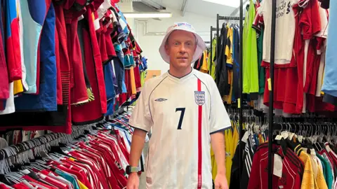 A man stands inside a shop aisle filled with hanging football shirts, wearing an England shirt and a hat.

