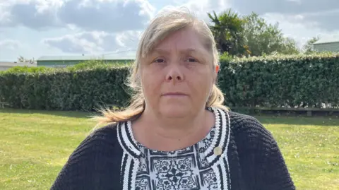 Qays Najm/BBC Julie Taylor has blonde hair, a black cardigan, and a stern expression. She is sitting outside, with grass and a hedge behind her. There are clouds above.