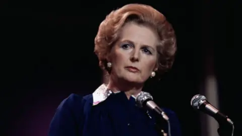 Margaret Thatcher has an auburn bob and large white pearl earrings. She is wearing a dark blue dress with a white collar and is standing in front of two black microphones on stands