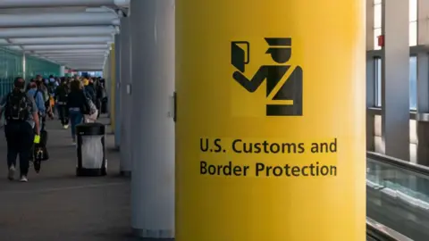 Inside airport. Lots of people walking with luggage on the left. On the right is a yellow pillar with the words US Customs and Border Protection written on it with a graphic of a male airport official looking at a passport.