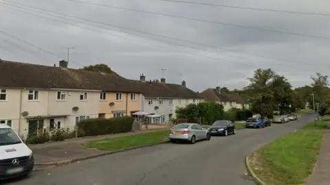 A row of terraced council houses on a residential road in Cirencester on a gloomy day. The houses are painted different light colours. There are cars parked on the road and some parasols outside one of the houses.