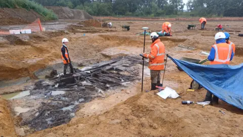 Cotswold Archaeology The wooden well discovery is shown in the foreground looking very wet, muddy and full of water, with one site worker stood within it and 5 other workers stood around it with wheelbarrows and other tools, some look to be analysing other holes. All workers are wearing high visibility clothing and hard hats.
