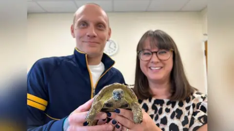 Sarah-May Buccieri/BBC A tortoise looking directly into the camera and looking content and happy with himself. He is being held up by Mr and Mrs Frost. Mr Frost is smiling and wearing a blue fleece and Mrs Frost is also smiling, wears glasses with brown hair, a fringe and a cheetah-print top.