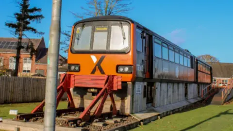 Northern An orange Pacer train is on a short section of track in a school playground which has green grass. The train has a black X branded on the front with a banner reading 'Newcastle'.