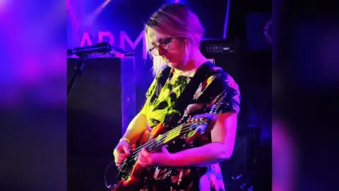 Jo Dale plays guitar onstage. She wears a patterned dress, and has short blonde hair and glasses. The stage lighting is purple, and in the background speakers and an amplifier can be seen.