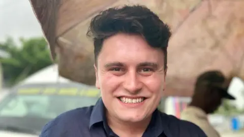 A young man is centre frame and smiling to camera. He has dark hair and is wearing a navy blue shirt.