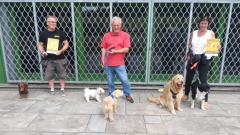 Three dog walkers with their dogs on leads holding Poo Bag Partners posters 