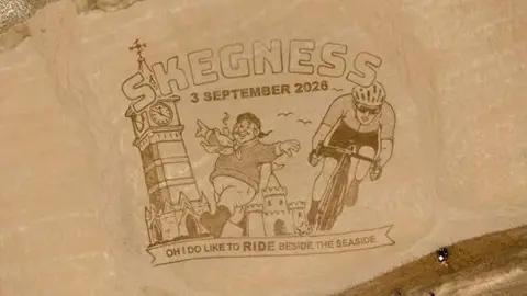 The sand art. It reads 'Skegness 3 September 2026' and depicts a clock tower, the Jolly Fisherman and a cyclist. At the bottom it says "Oh I do like to ride beside the seaside" in capital letters. 