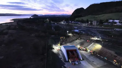 Network Rail A general view of a concrete tunnel next to a road, with heavy machinery around t. There is a body of water and hills in the background at dusk. 
