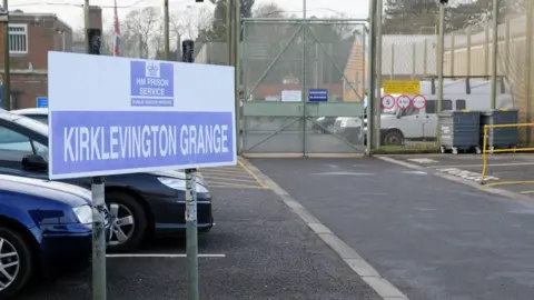 A blue and white sign with the HM Prison Service logo and, in capital letters, the name Kirklevington Grange. It stands on two grey poles and sits in a car park in front of a very high, greenish-grey mesh fence and metal gates topped with barbed wire. Low, brick buildings can be seen beyond the gates and fence.