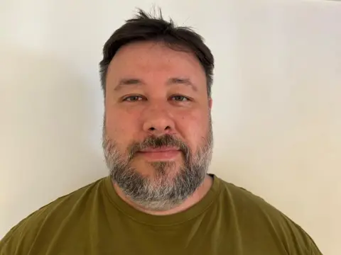 Wesley Yin-Poole A man with short black hair and a neatly-trimmed beard stands in front of a blank cream background. He's wearing an olive-coloured t-shirt.