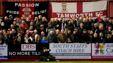 Tamworth Football Club Tamworth fans in the stands. There are sponsorship boards around them and flags in the background. 