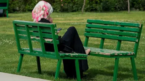 A woman in a headscarf on a green bench in the park. She has a pink and white floral scarf on and a black dress on.