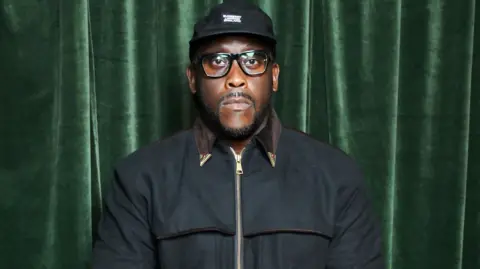 Getty Images Kenny Allstar pictured in December 2024. He wears a black cap with a black zipped-up jacket. He has black rimmed square glasses and a trimmed moustache and beard. He's pictured with a serious expression in front of a green velvet curtain. 