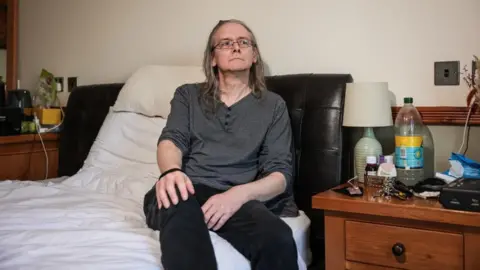LDRS Martin Atkinson, a middle-aged man with long grey hair, sits on a bed looking to the side of the camera. He wears black jeans and a long-sleeved grey shirt. 