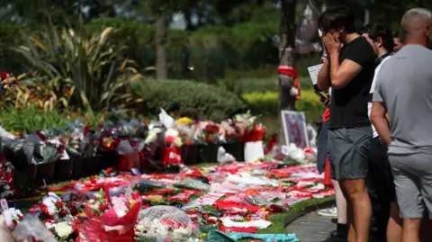 A crowd of people, including a man with his face in his hands, stand on the edge of a grassy area blanketed in flowers, football shirts, scarves and balloons