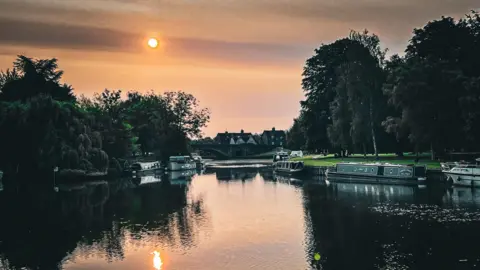 Becca C A river with several narrowboats and a low sun reflecting on the water.