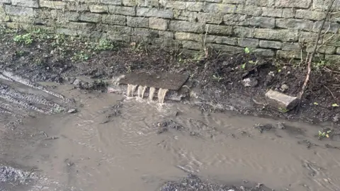 Contributed In the centre of the shot is a drain which isn't fully closed,  with brown, discoloured water flowing out on to the road, which is also covered in the same colour water. 