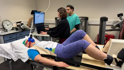 Volunteer Julie Smith lies on bench in the foreground. She wears sports gear and a blue mask with a plastic tube. Her feet are strapped into a horizontal stepping fitness machine. Behind her Dr Jo Mallinson (in a black top) and Dr Abhishek Sheth (in green scrubs) monitor a screen and laptop