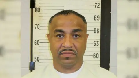 A mugshot of Tony Carruthers. He wears a beige shirt, has a mustache, and stares straight ahead. 